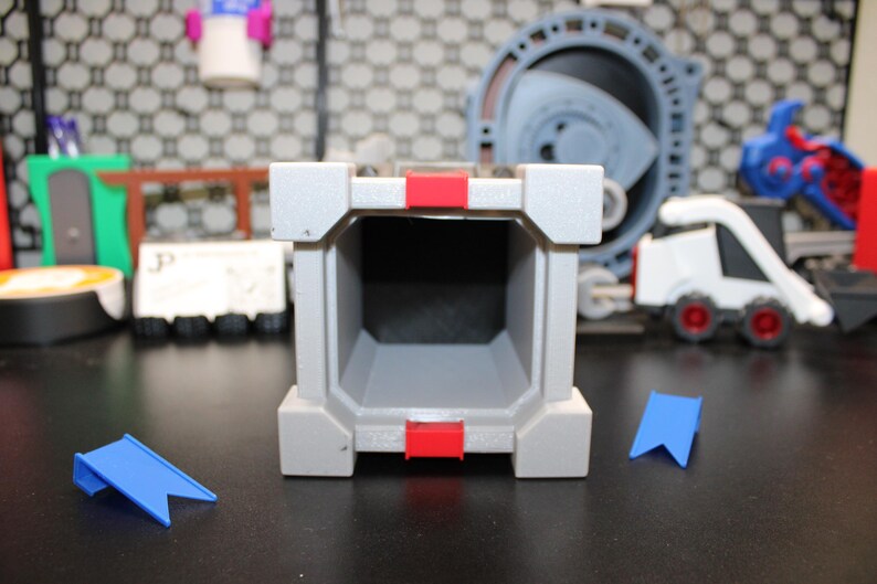 May include: A grey, rectangular toy structure with red accents and a hollow interior. Two blue, flag-shaped pieces are in front of the structure. Other toys are in the background, including a white vehicle with red wheels.