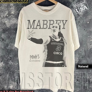 Può includere: Una t-shirt color naturale con una stampa grafica nera di una giocatrice di basket che indossa una maglia di Toronto con il numero 3. Il testo MABREY è stampato sopra la giocatrice e MM#3 53 POINTS sotto il braccio della giocatrice.
