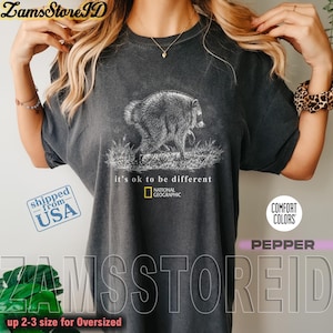 May include: A charcoal gray short-sleeve t-shirt featuring a white line-art illustration of a raccoon walking through grass. Below the graphic, the text reads "it's ok to be different" followed by the National Geographic logo in yellow and white.