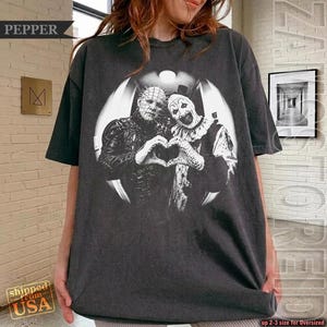 May include: A charcoal gray short-sleeve t-shirt featuring a black and white graphic print of Pinhead and Art the Clown forming a heart shape with their hands. The design is set within a circular frame on the front of the garment.
