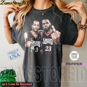 May include: A charcoal gray short-sleeve t-shirt featuring a graphic print of two basketball players in jerseys with the numbers 3 and 23. The text "SIXERS" is printed on the chest. The shirt has a vintage-style wash and a crew neckline.