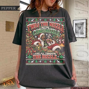 May include: A charcoal grey t-shirt featuring a graphic print of a person in a Santa suit holding a mug. The text reads: TWAS THE NIZZLE BEFORE CHRISMIZZLE AND ALL THROUGH THE HIZZLE. The design includes Christmas trees, presents, and cannabis leaves.
