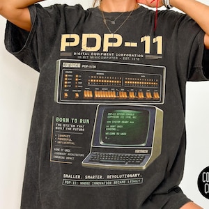 May include: Dark gray t-shirt featuring a vintage computer graphic with the text "PDP-11" and "Digital Equipment Corporation." The design includes a computer console illustration and the phrase "Born to Run." The shirt has a retro aesthetic.