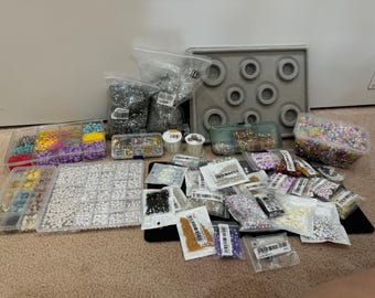 May include: A variety of colorful beads and sequins in small plastic containers, organized on a table. There are also several bags of beads and a gray tray with circular indentations.