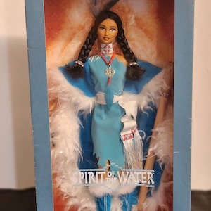 Könnte beinhalten: Eine limitierte Barbie-Puppe in einem blauen Kleid mit weißem Pelzbesatz. Die Puppe trägt eine Perlenkette und hat lange schwarze Zöpfe. Die Schachtel zeigt den Text "Spirit of the Water" und "Barbie Collectibles".