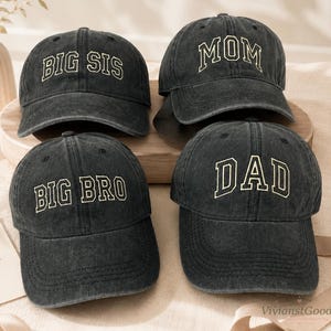 Puede incluir: Cuatro gorras de béisbol gris carbón con un acabado desgastado. Cada gorra presenta letras bordadas en color crema claro. Las gorras llevan los textos BIG SIS, BIG BRO, MOM y DAD en una tipografía llamativa de estilo universitario.