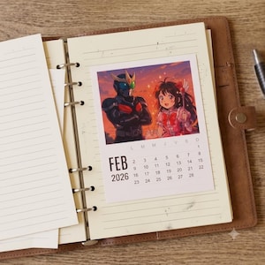 Puede incluir: Una agenda de anillas de cuero marrón con papel rayado y una página de calendario de febrero de 2026. El calendario presenta una ilustración estilo anime de una figura con armadura negra y una chica de pelo oscuro sosteniendo un bastón.