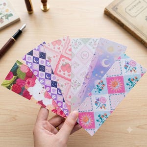 Puede incluir: Un conjunto de seis marcapáginas rectangulares de papel abanicados, con diversos patrones que incluyen frambuesas rojas con un animal blanco, rombos morados con lunas, fresas rosas, un enrejado floral blanco, degradados celestiales y soles con rosas.