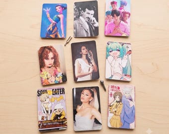 Puede incluir: Ocho tarjetas coleccionables rectangulares con imágenes diversas, incluyendo personajes de anime, un retrato en blanco y negro de Nikola Tesla, una mujer con vestido blanco, una estrella del pop y una portada de revista Soul Eater, con un diapasón.