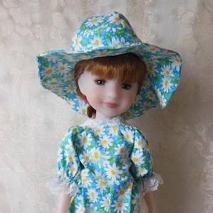 May include: A doll wearing a blue jumpsuit and matching hat, both adorned with a white daisy pattern. The doll has fair skin, red hair, and white shoes. The outfit features lace trim on the sleeves and neckline.