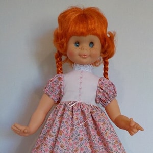 May include: A red-haired doll wearing a pink floral dress with a white bodice and black shoes. The doll has blue eyes and a sweet smile.
