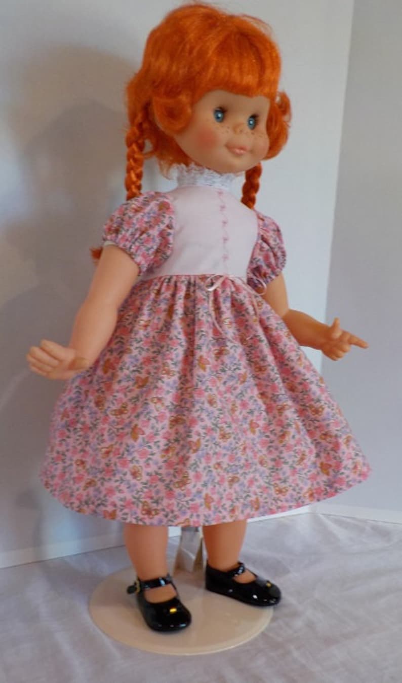 May include: A red-haired doll wearing a pink and white floral dress with black shoes. The doll has braids and blue eyes.