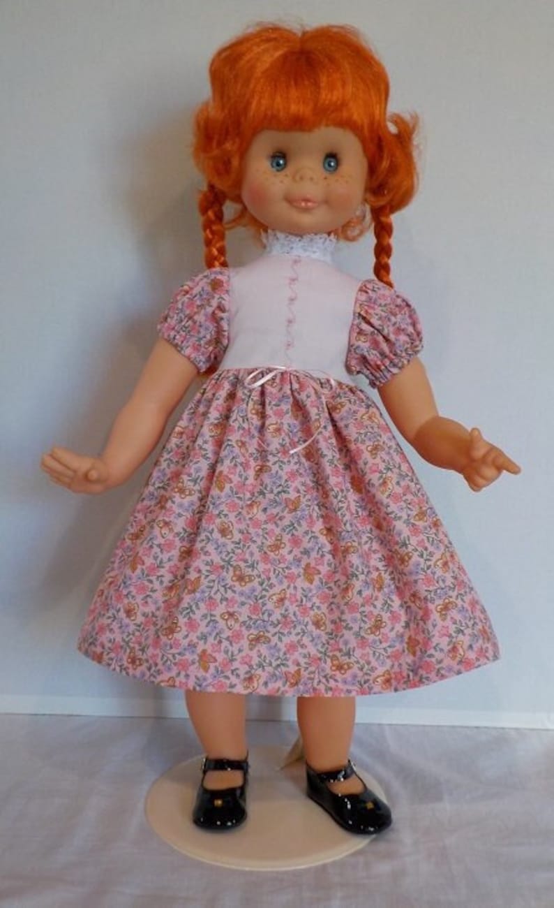 May include: A red-haired doll wearing a pink floral dress with a white bodice and black shoes. The doll has blue eyes and a sweet smile.