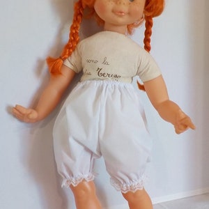 May include: A red-haired doll with blue eyes and freckles wearing a white shirt with the text "como la cerveza" and white bloomers with lace trim.
