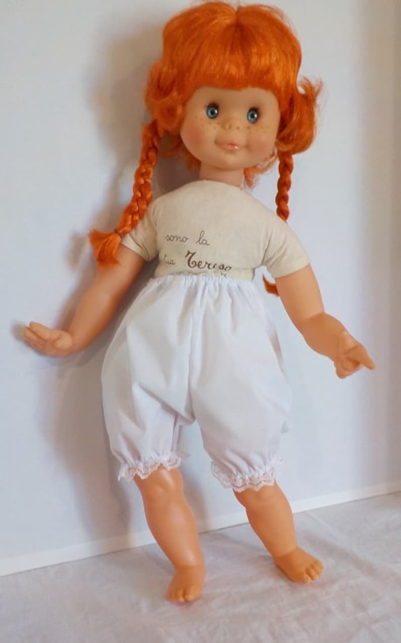May include: A red-haired doll with blue eyes and freckles wearing a white shirt with the text "como la cerveza" and white bloomers with lace trim.