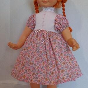 May include: A red-haired doll wearing a pink dress with a floral pattern and black shoes. The doll has blue eyes and a white lace collar.