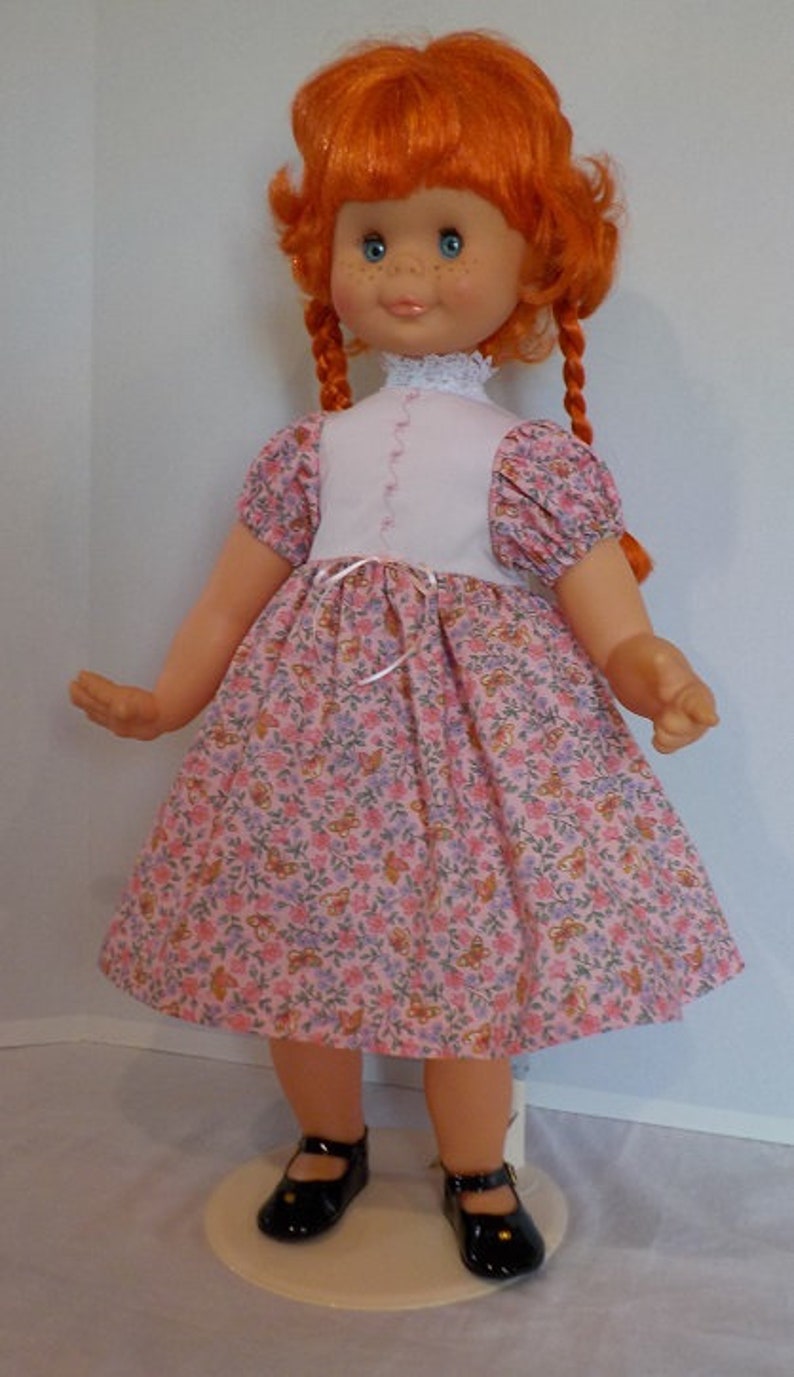May include: A red-haired doll wearing a pink dress with a floral pattern and black shoes. The doll has blue eyes and a white lace collar.