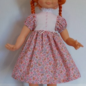 May include: A red-haired doll wearing a pink and white dress with a floral pattern. The doll has blue eyes and is wearing black shoes.