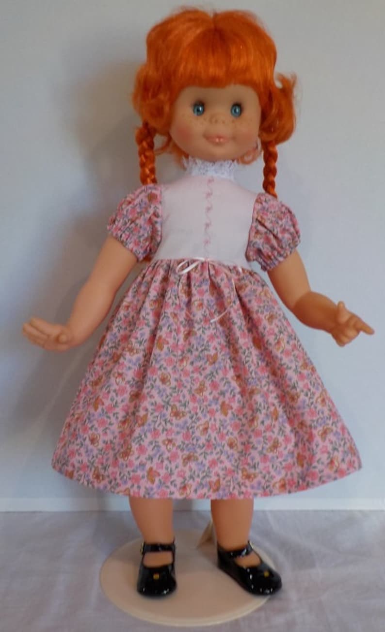 May include: A red-haired doll wearing a pink and white dress with a floral pattern. The doll has blue eyes and is wearing black shoes.