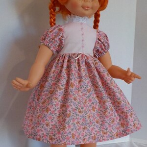 May include: A red-haired doll wearing a pink dress with a white bodice and floral print. The doll has blue eyes and freckles and is wearing black shoes.