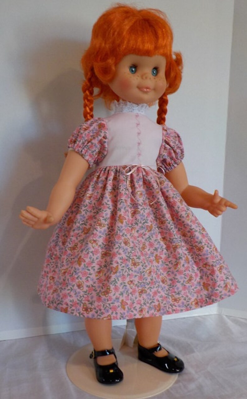 May include: A red-haired doll wearing a pink dress with a white bodice and floral print. The doll has blue eyes and freckles and is wearing black shoes.
