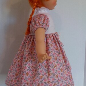 May include: A doll with red hair wearing a pink and white dress with a floral pattern. The doll is wearing black shoes and is standing on a white stand.