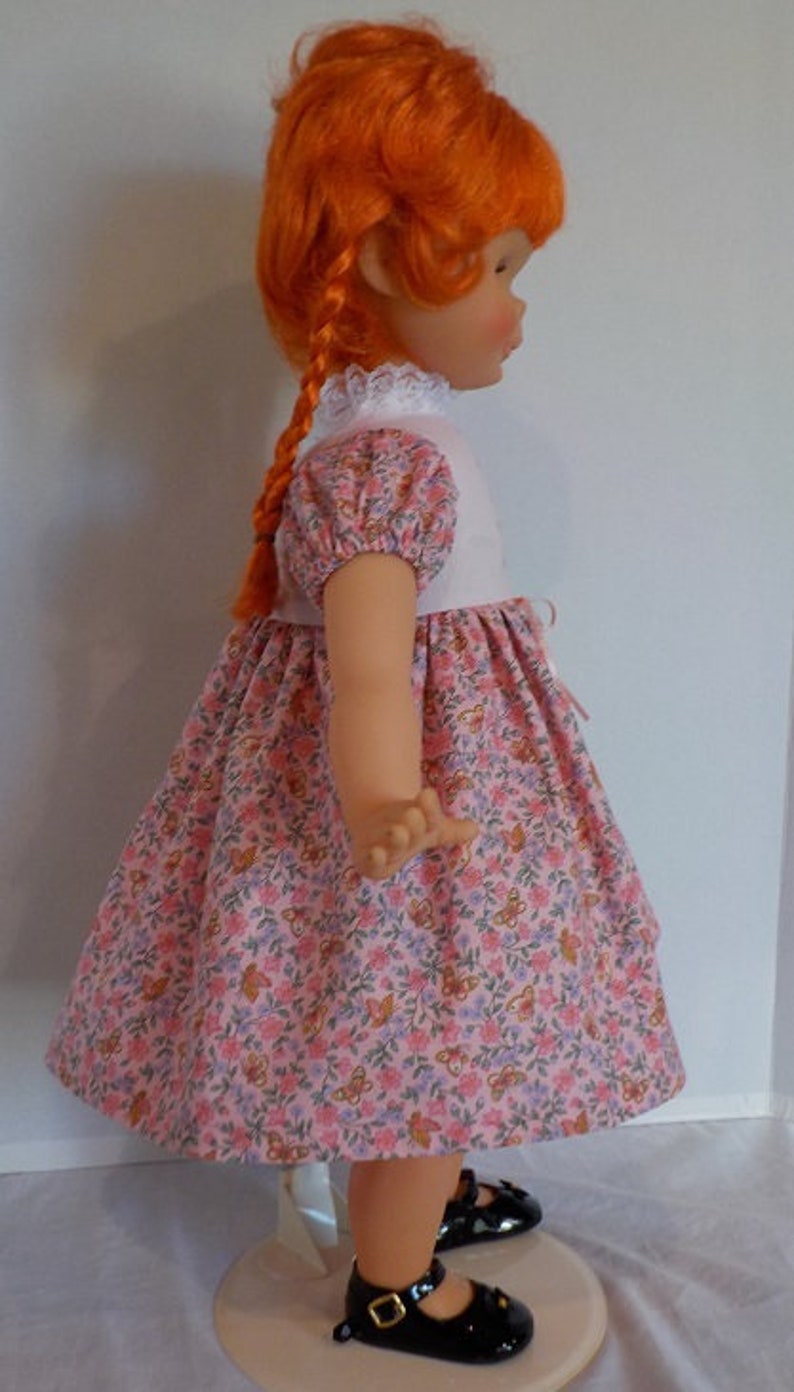 May include: A doll with red hair wearing a pink and white dress with a floral pattern. The doll is wearing black shoes and is standing on a white stand.