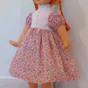 May include: A red-haired doll wearing a pink dress with a white bodice and floral print. The doll has blue eyes and is wearing black shoes.