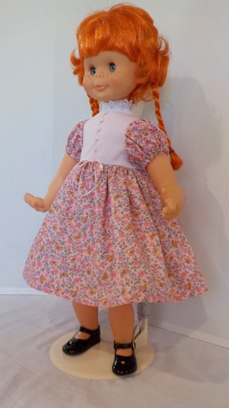May include: A red-haired doll wearing a pink dress with a white bodice and floral print. The doll has blue eyes and is wearing black shoes.
