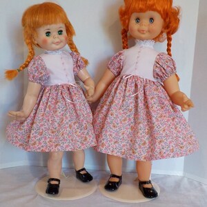 May include: Two red-haired dolls wearing matching pink and white dresses with floral patterns. The dolls have pigtails and are wearing black shoes.