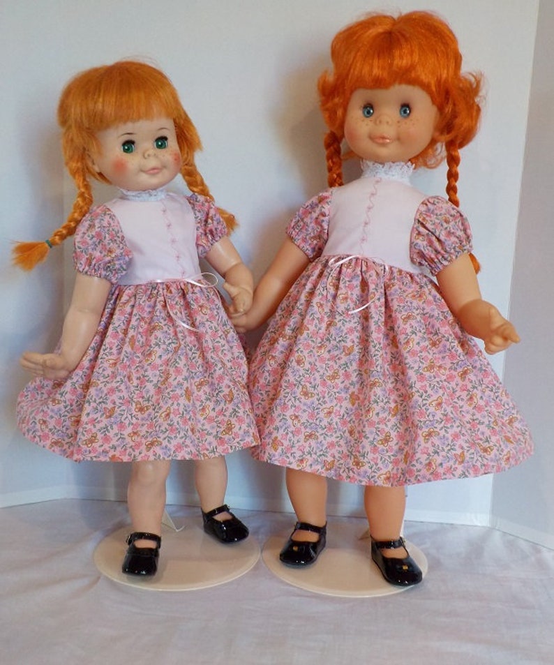 May include: Two red-haired dolls wearing matching pink and white dresses with floral patterns. The dolls have pigtails and are wearing black shoes.