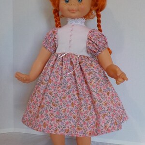 May include: A red-haired doll wearing a pink and white dress with a floral pattern. The doll has blue eyes and is wearing black shoes.