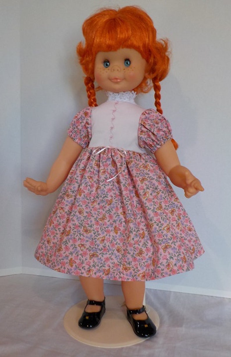 May include: A red-haired doll wearing a pink and white dress with a floral pattern. The doll has blue eyes and is wearing black shoes.