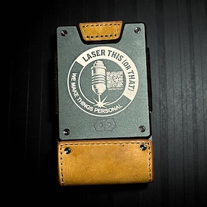 May include: A gray metal wallet with tan leather accents. The wallet features a circular graphic with the text "LASER THIS (OR THAT)" and "WE MAKE THINGS PERSONAL." The wallet is rectangular with rounded corners and visible screws.