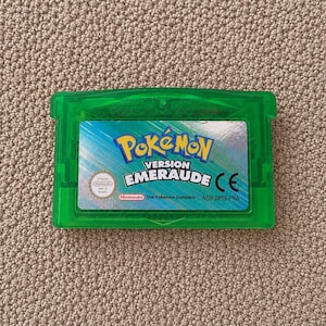 May include: A translucent green Game Boy Advance video game cartridge titled Pokémon Version Emeraude. The label features the Pokémon logo, the game title, a Nintendo seal of quality, and the text The Pokémon Company AGB-BPEF-FRA.