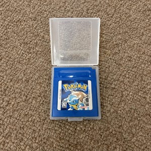 May include: A blue Nintendo Game Boy video game cartridge for Pokémon Blue Version, housed in a clear plastic protective case. The label features the Pokémon Blastoise, the Pokémon logo, an ESRB E rating, and the text DMG-APEE-USA.