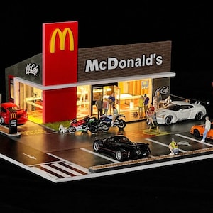 May include: A detailed miniature McDonald's diorama, featuring the restaurant's facade with the golden arches. Includes toy cars, motorcycles, and figures, with a drive-thru sign. Set against a black backdrop.