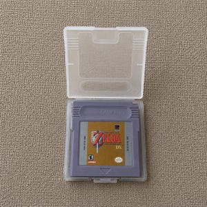 May include: A purple Game Boy video game cartridge for The Legend of Zelda: Link's Awakening DX, featuring a gold label with the game title, a sword and shield graphic, and an E rating, housed in a clear plastic protective storage case.