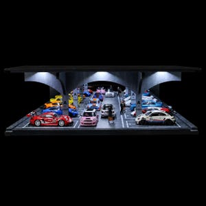 Op de afbeelding: Een miniatuur parkeergarage vitrine met diverse gegoten speelgoedauto's in verschillende kleuren, waaronder rood, roze, blauw en wit. De constructie is voorzien van ledverlichting, steunpilaren gemarkeerd met de letter A en geschilderde parkeerlijnen.