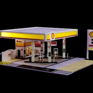 Può includere: Modello in miniatura di un distributore di benzina Shell, con tettoia gialla e rossa, pompe di benzina e un negozio. Il modello include un cartello Shell e un display dei prezzi, su sfondo nero.