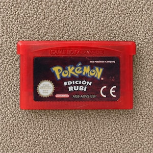 May include: A red Game Boy Advance video game cartridge for Pokémon Edición Rubí. The label features the Pokémon logo, the text Edición Rubí, The Pokémon Company, Nintendo, a circular seal of quality, the CE mark, and the code AGB-AXVS-ESP.