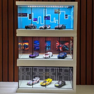 May include: A three-tier display case with integrated LED lighting, featuring glass panels and individual compartments. Each level houses three miniature toy cars against distinct backdrops, including a blue skyline, a red wall, and a white line design.