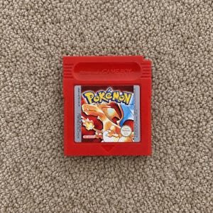以下が含まれることがあります： 赤色の任天堂ゲームボーイ用ビデオゲームカートリッジ、ポケットモンスター赤バージョン。ラベルには、翼を広げた炎タイプのポケモン、リザードンと、その尻尾の炎のイラストが描かれています。ラベルには「Pokémon」および「Nintendo」の文字が記されています。