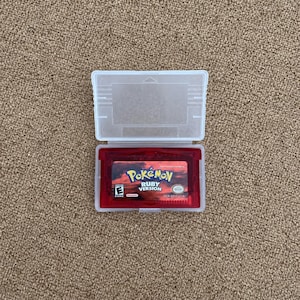 Puede incluir: Un cartucho de videojuego para Game Boy Advance de color rojo translúcido titulado Pokémon Ruby Version, con una etiqueta que muestra un cielo al atardecer, dentro de una funda protectora de plástico transparente abierta hacia arriba.