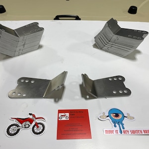 May include: A set of 20 silver metal key switch brackets. The brackets are stacked in two piles of 10. There are two individual brackets in the center of the image. The image also includes a business card with the text "Norm & Amy Dahl" and a sticker with the text "Move It Key Switch Bracket".