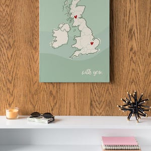 Può includere: Una stampa su tela verde con una mappa del Regno Unito. Due cuori rossi segnano le posizioni sulla mappa. Il testo "with you." è stampato sotto la mappa.