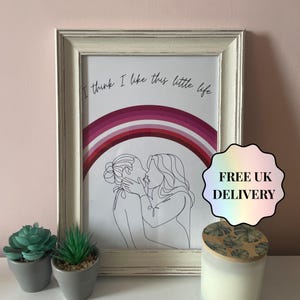 Könnte beinhalten: Gerahmter Kunstdruck mit einer Strichzeichnung von zwei sich küssenden Personen, einem Regenbogen und dem Text "I think I like this little life". Der Rahmen ist cremefarben. Eine Kerze und kleine Topfpflanzen sind im Vordergrund.