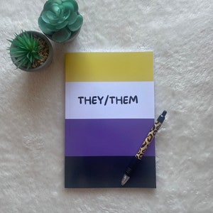 Peut inclure: Un carnet avec une couverture jaune, violette et noire. La couverture porte l'inscription "THEY/THEM". Un stylo à imprimé léopard est posé sur le carnet.