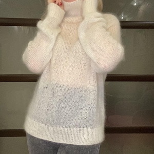 May include: A woman wearing a cream-colored, oversized turtleneck sweater. The sweater has a loose, textured knit. She is also wearing gray leggings. The background is a dark, neutral color.