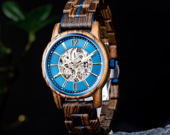 May include: A wooden wristwatch with a blue face and a visible mechanical movement. The watch has a wooden band with blue accents. The watch face displays the numbers 12 and 6. The watch is displayed on a dark, textured surface.